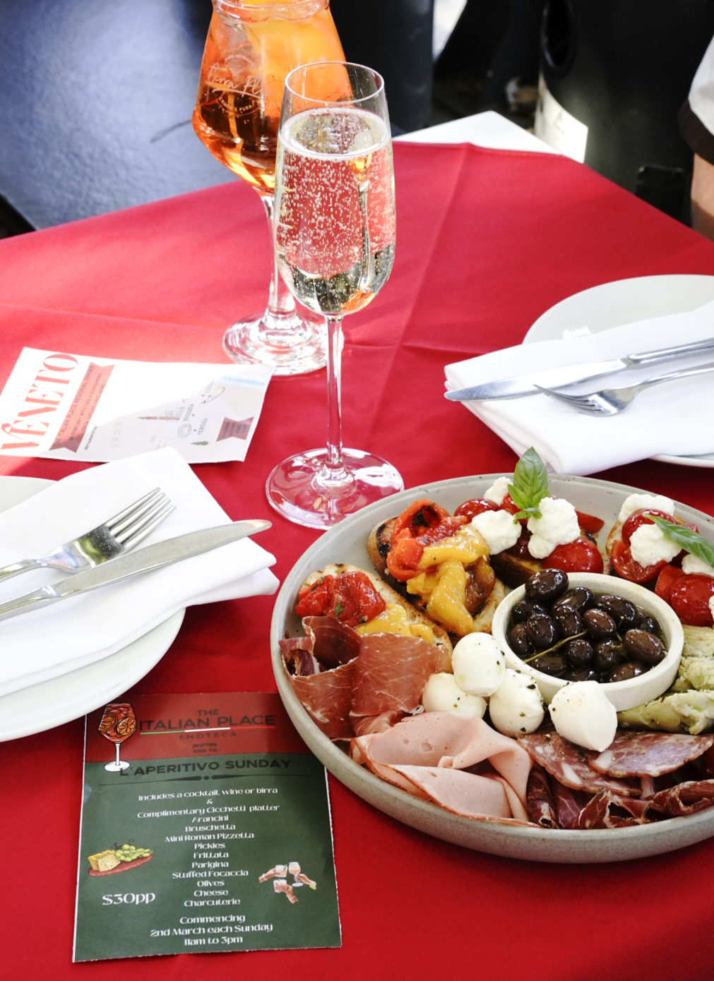 A plate of antipasto and snacks on a red tablecloth with a glass of bubbles and an Aperol Spritz.