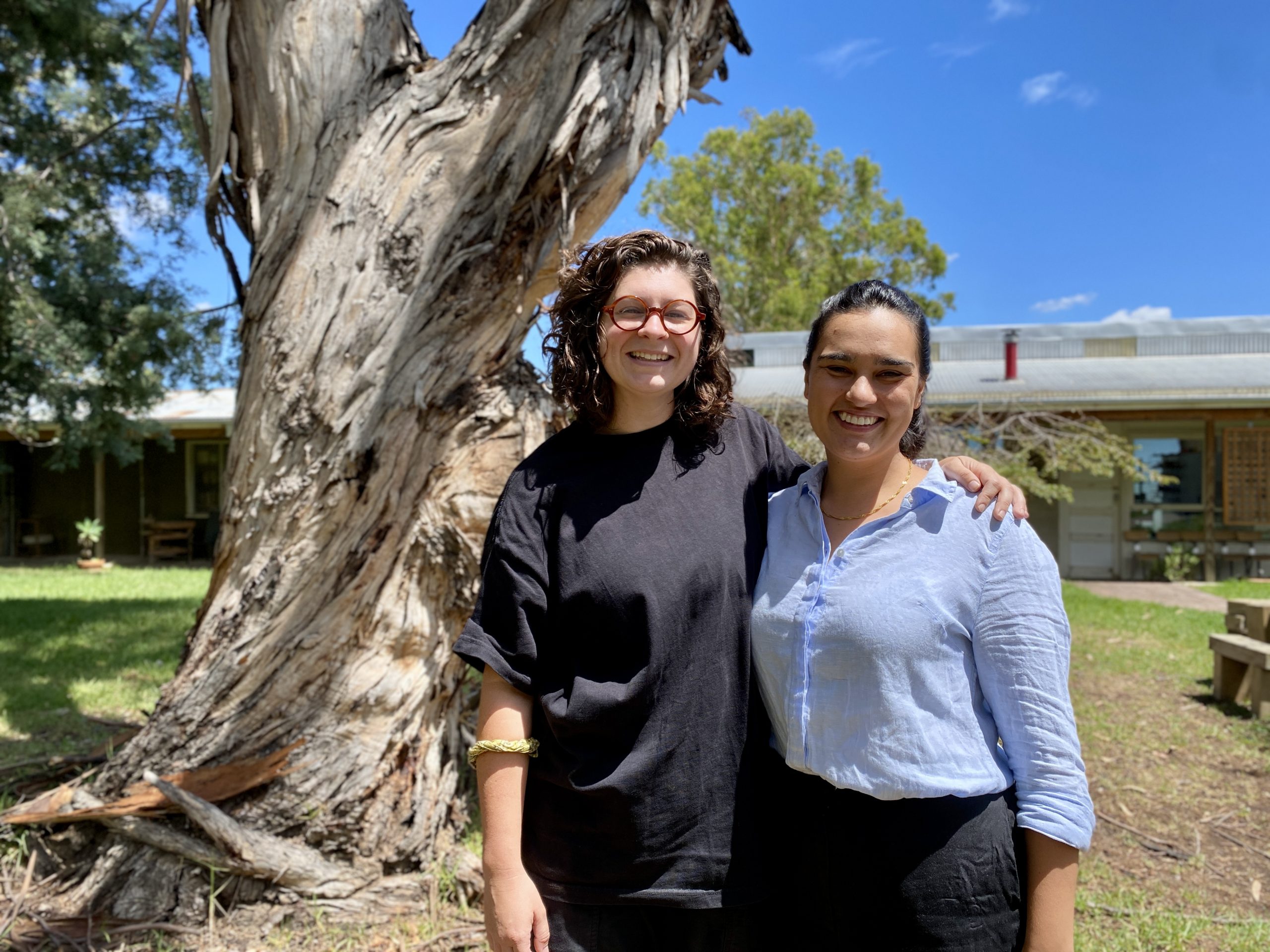 Two women stand in front of a large gum tree on a sunny day. 