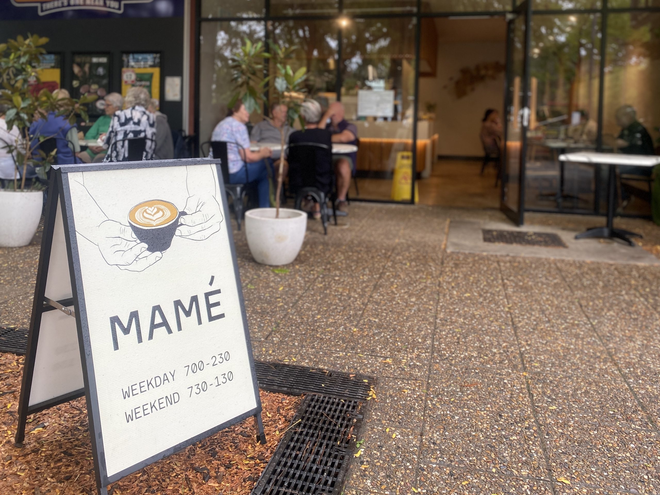 A-frame sign reading Mame with a picture of a cup of coffee held in hands.