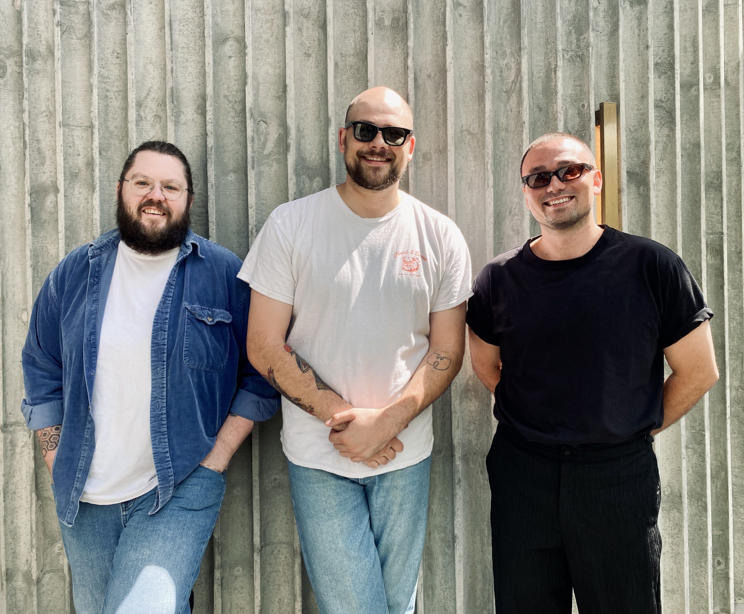 Three men stand against a textured concrete wall.