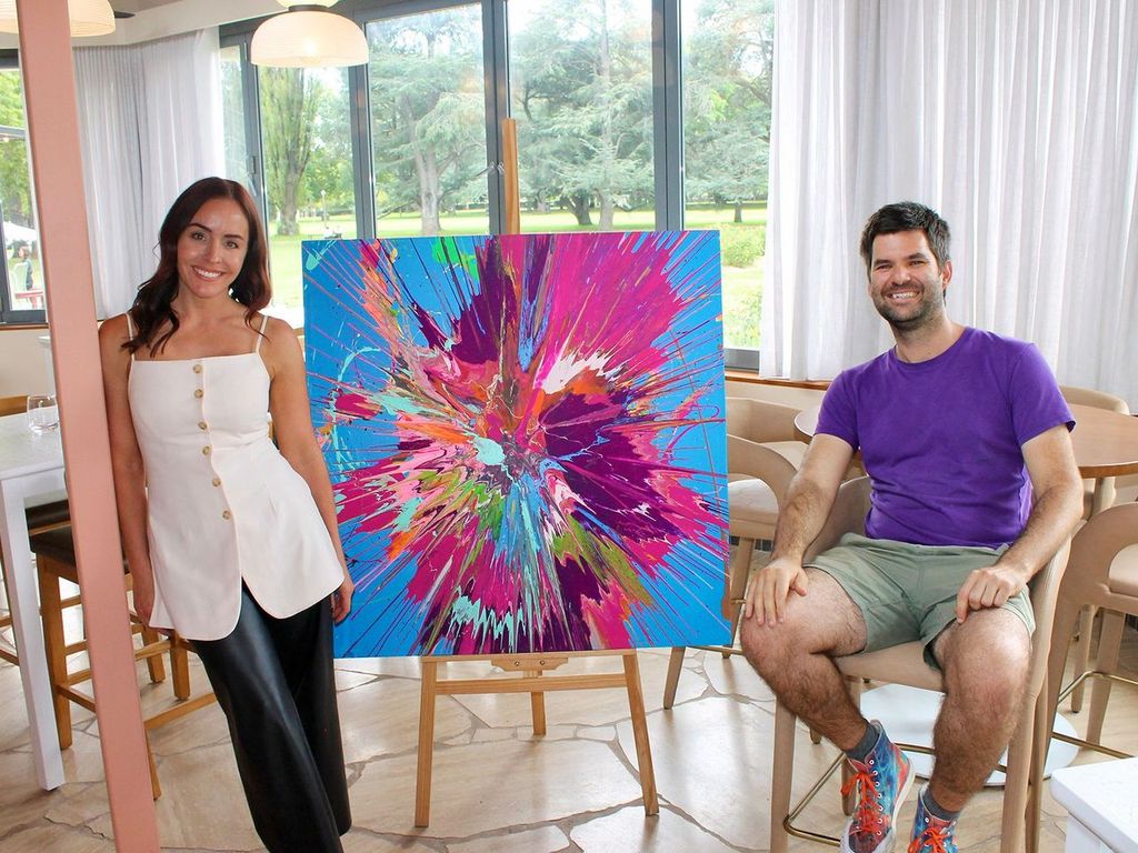 A woman in a white top stands to the left of a vibrant pink and blue painting, on the right side sits a man in shorts and a purple t-shirt.