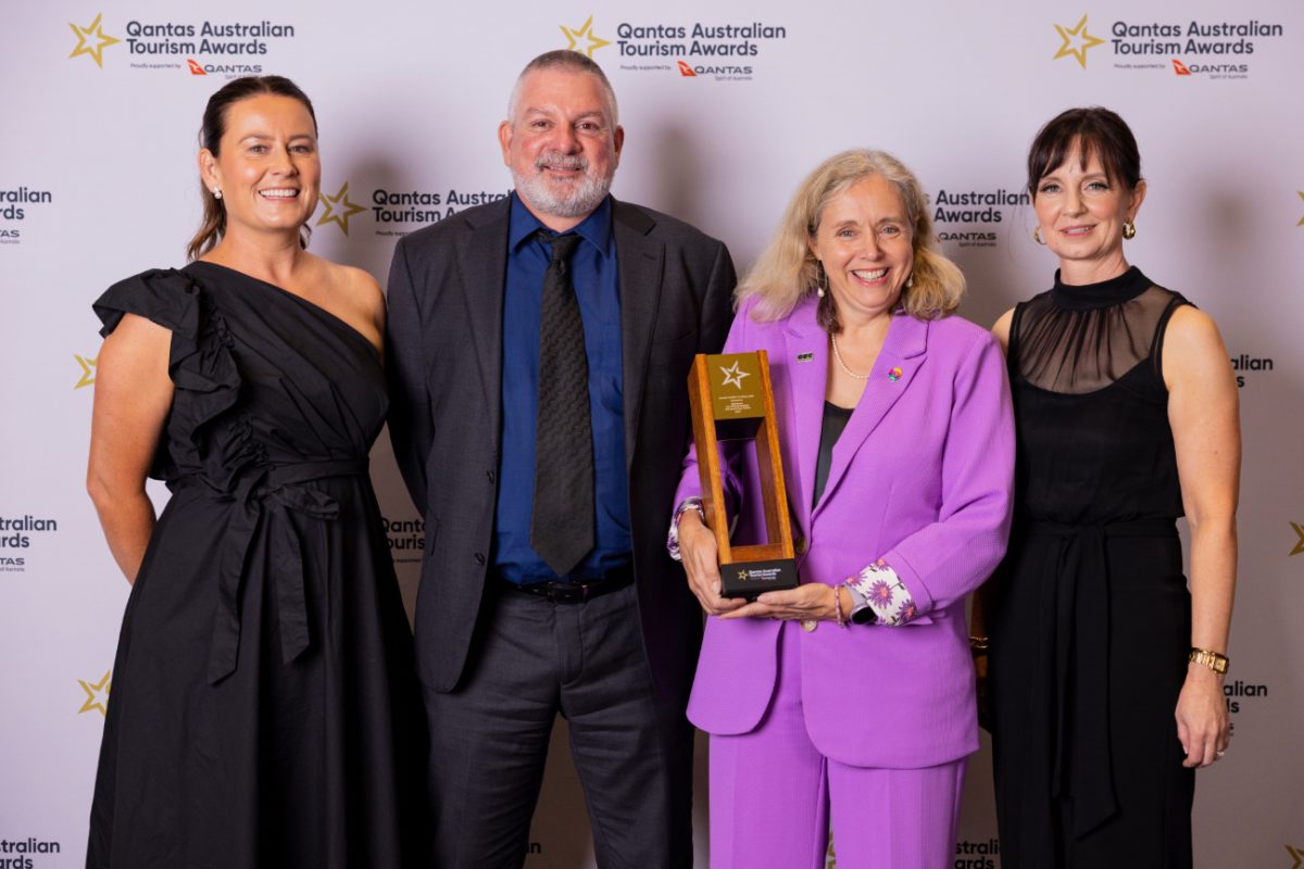 Three women and a man in formal wear standing together, one woman holding a trophy