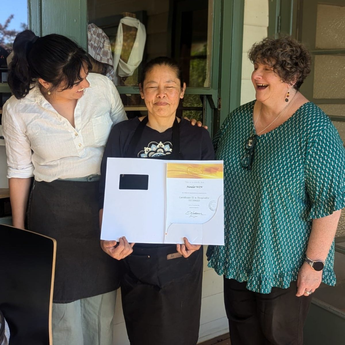 An older woman smiles proudly with her eyes closed in the centre holding a certificate which reads "Certificate III in Hospitality". Two women stand on either side of her smiling.