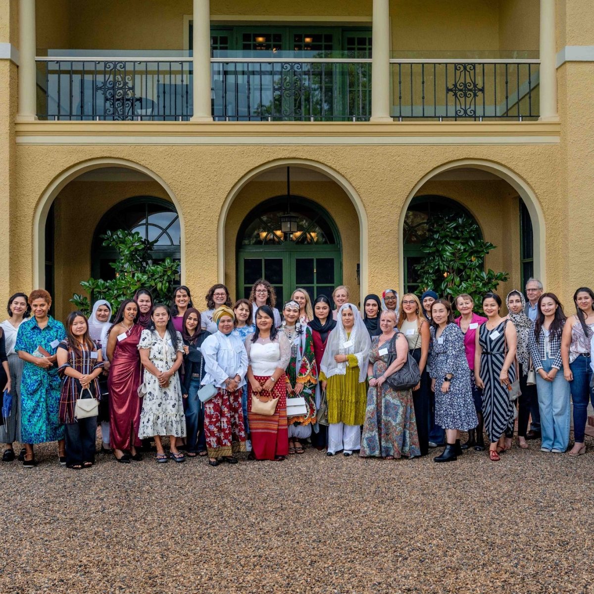 A group of women dressed in different traditional clothes from around the world gather for a group photo in front of a large yellow building with arches.