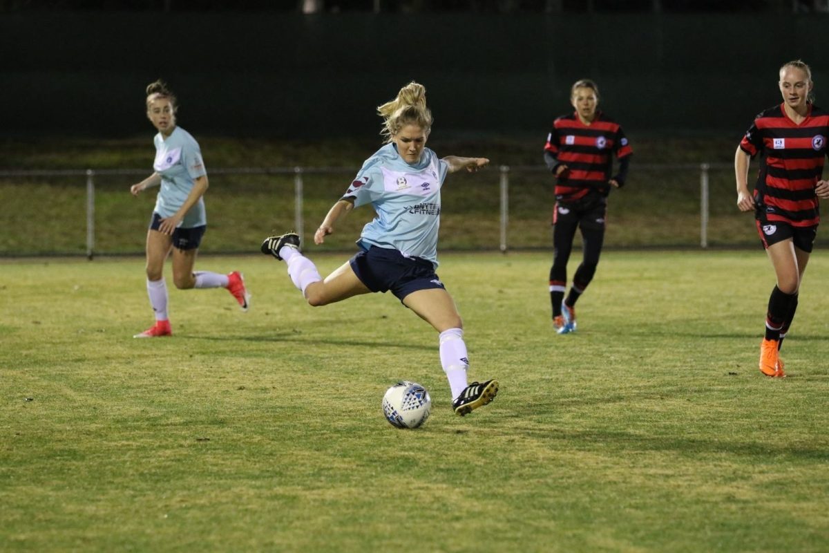 An action shot of a female soccer playing in a blue uniform kicking the ball.