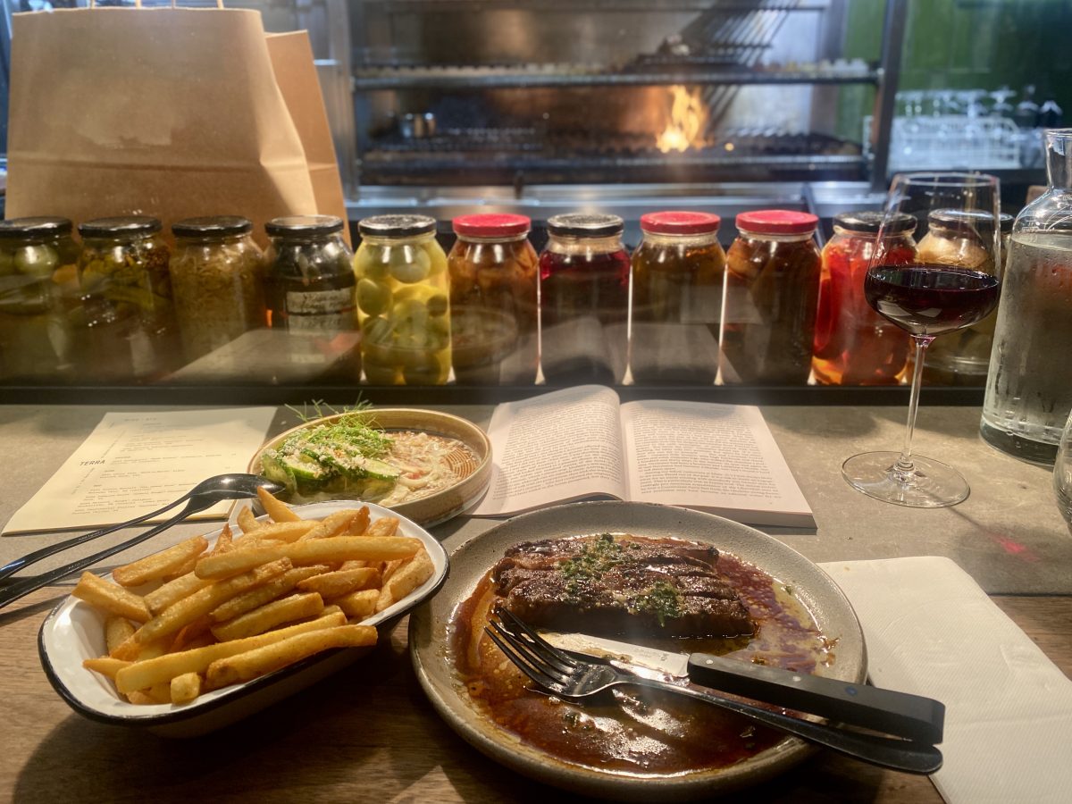 A view of a partially eaten meal: steak, chips and a cucumber salad with a glass of red wine, an open book and a view into an open kitchen with flames in the grill.