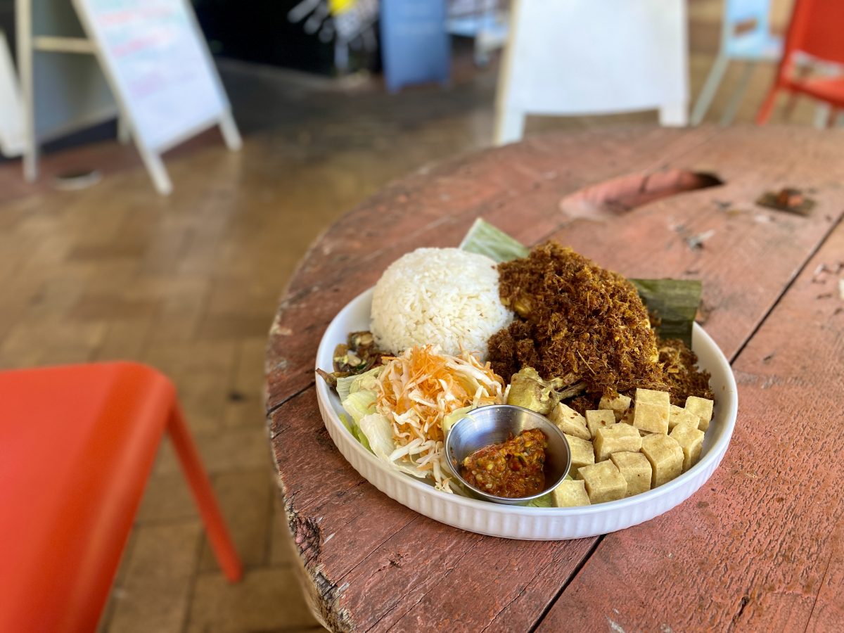 A plate on the edge of a rustic looking outdoor table. The plate has a range of ingredients on it: a mound of rice, a piece of chicken, cubes of tofu, vegetables and chilli sauce in a small metal dish.