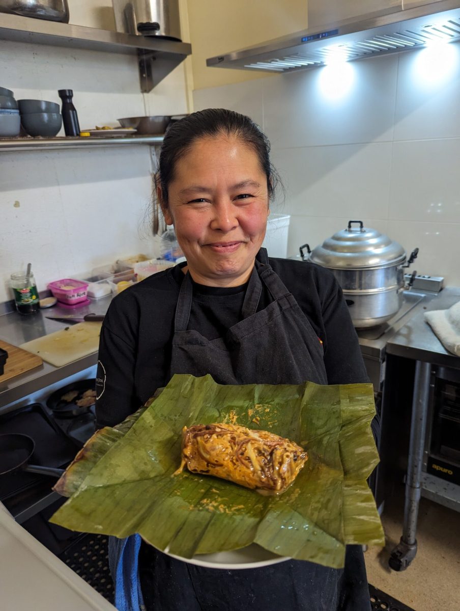 An Asian woman wearing a black apron stands in a kitchen and holds a piece of banana leaf which has been unwrapped to display a dish of banana blossom inside.