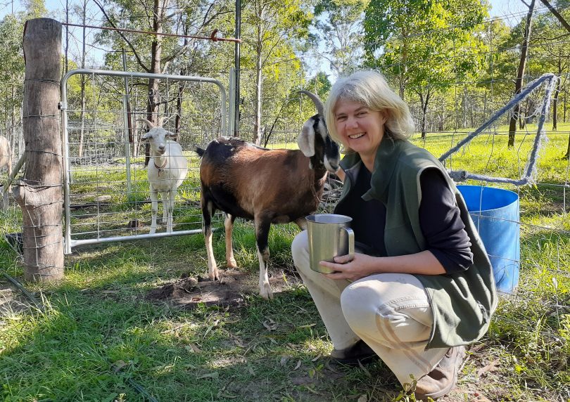 Maike Quellenburg of Bally Park Goats Milk Soap crouching on her farm in front of brown goat while holding jug of goats' milk.