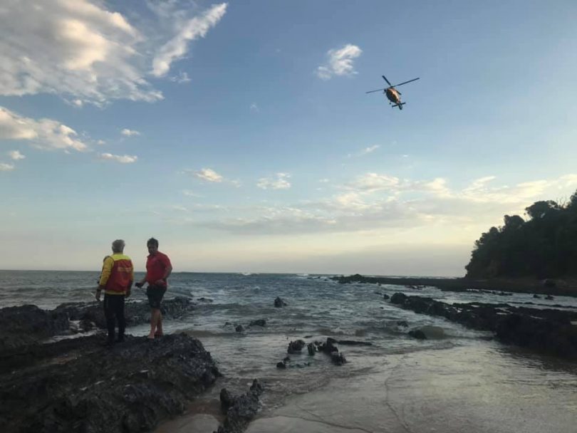 Two Surf Lifesavers stand on rocks at Richmond Beach while the Westpac Rescue Helicopter flies overhead.