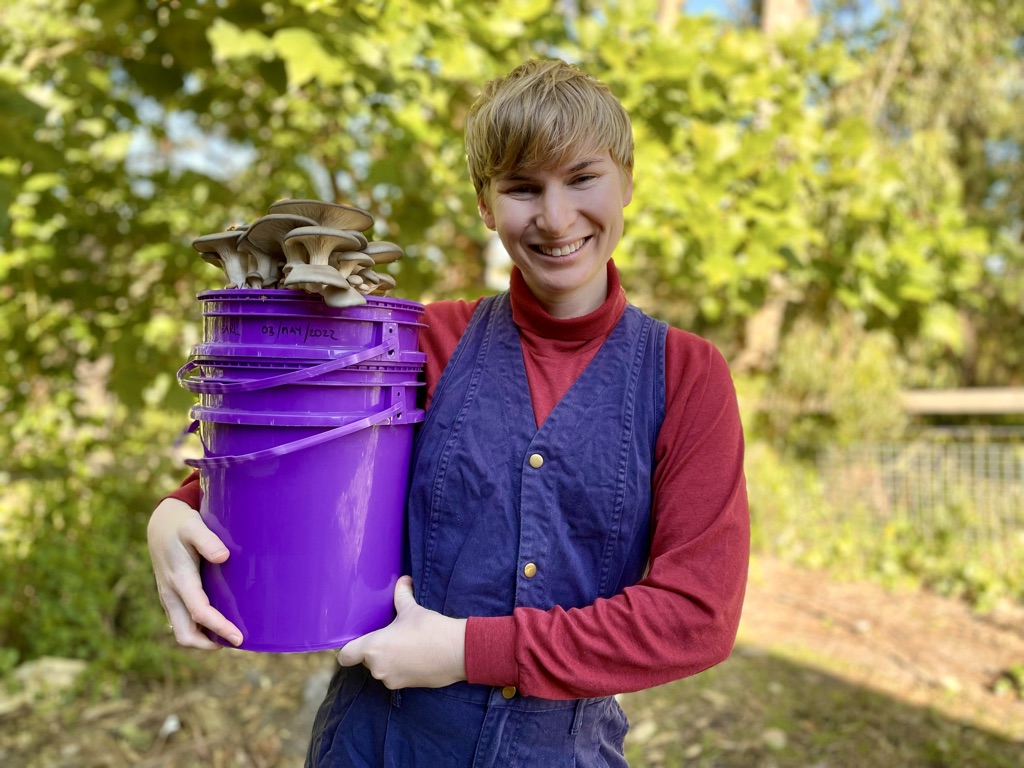Marita smiles and wears blue overalls, she is holding a large purple bucket with mushrooms growing out of the top.