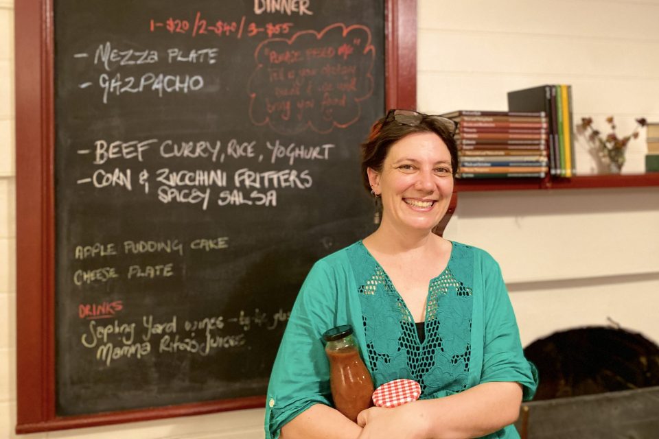 RA woman holding preserves in bottles stands in front of a restaurant blackboard