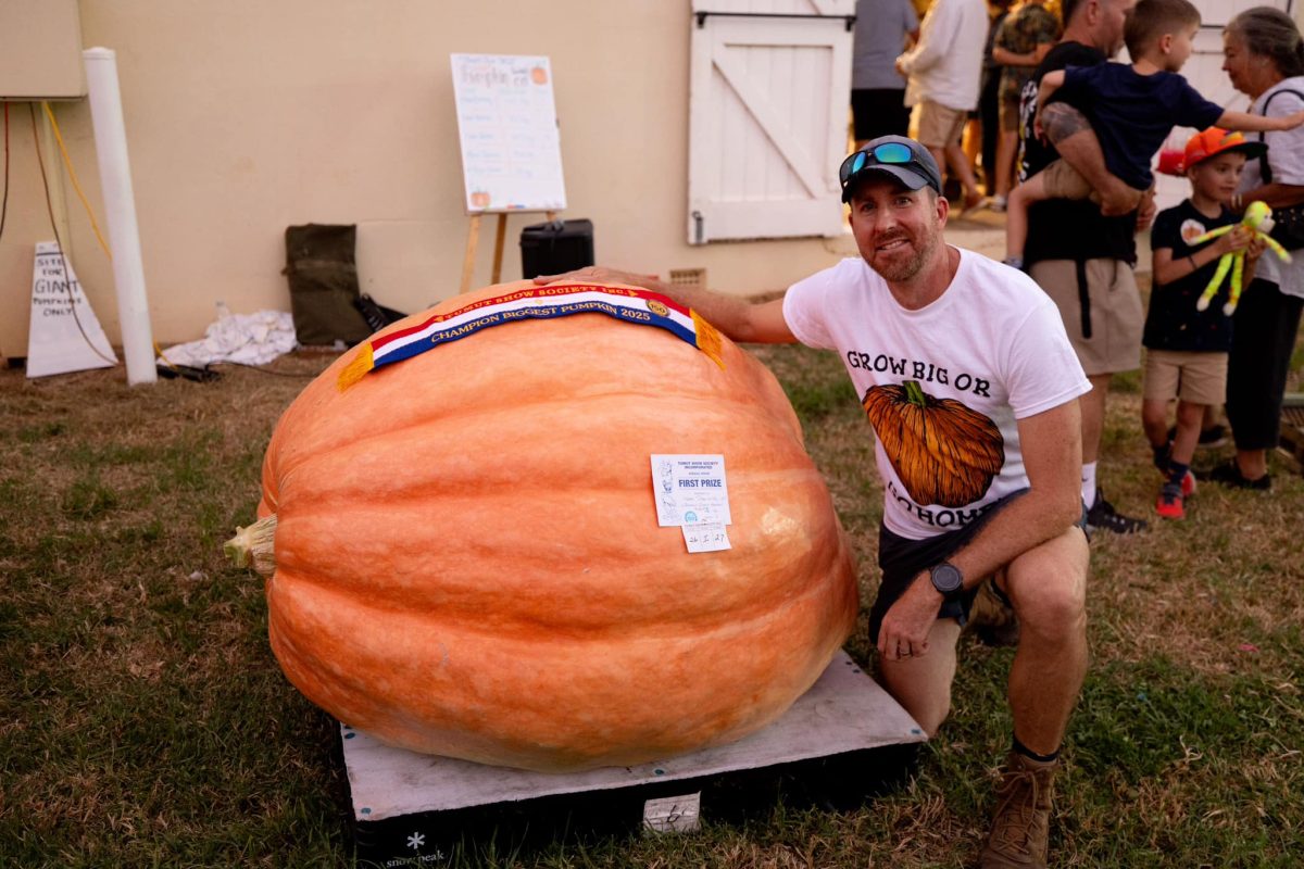 A man kneels next to a giant pumpkin that's draped with winning ribbons